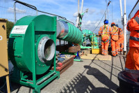 VENTEX Centrifugal Fan 450S with ducting entering a small hole on the platform nearby. Workers in orange overalls stand next to the hole with water in the background