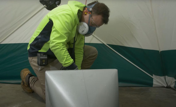 Worker in a respirator and hi vis kneeling beside ductwork inside an enclosure