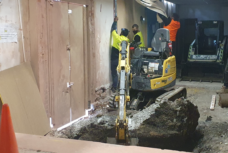 Excavator breaking out a floor inside a building with workers alongside