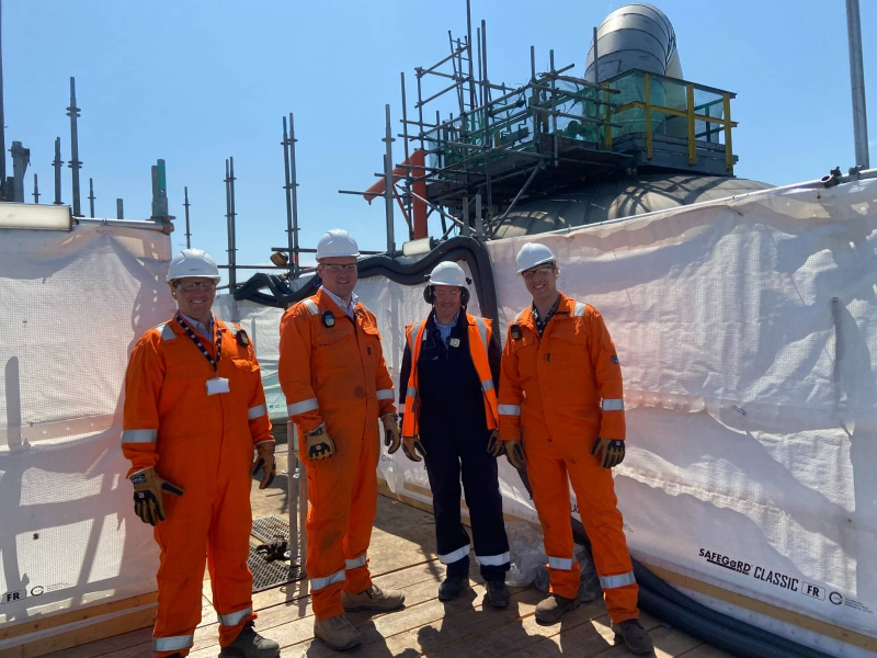 Four workers in coveralls in front of a sheeted refinery structure