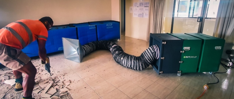 Worker removing floor tiles beside a dust containment barrier and RVT dust filtration unit